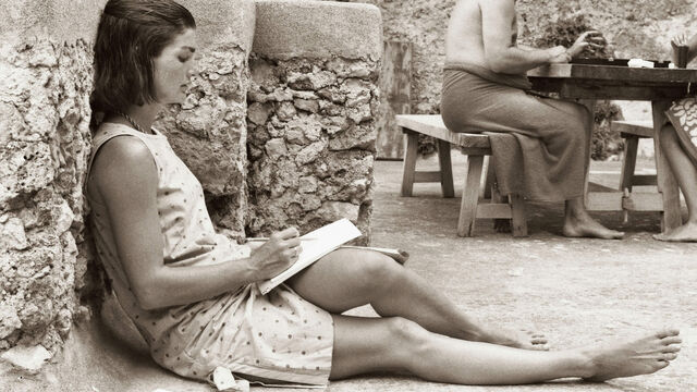 Black and white photo of Jackie Kennedy sitting down with a notebook on her lap